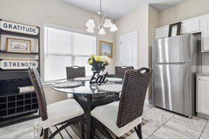 A modern kitchen with a dining table surrounded by wicker chairs, a stainless steel refrigerator, and a window with blinds. - Furnished 3BR Home Near Baylor Scott & White, Monthly Stays (Temple)