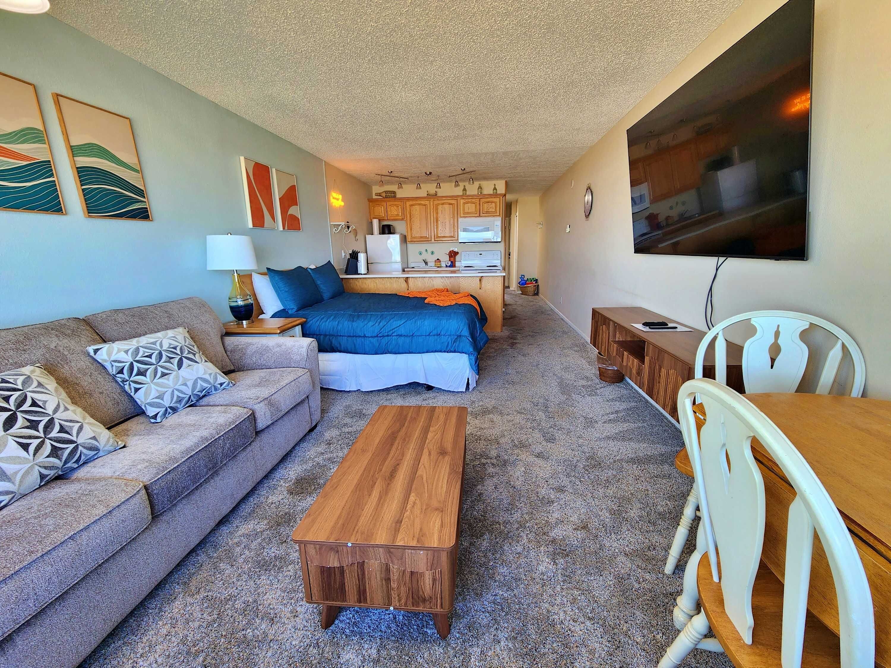 A compact living space with a sofa, a bed, a wooden table, and a kitchenette in the background.