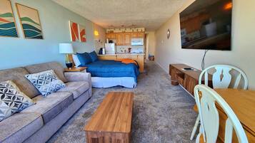 A compact living space with a sofa, a bed, a wooden table, and a kitchenette in the background.