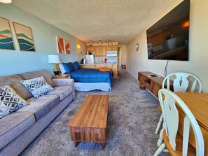 A compact living space with a sofa, a bed, a wooden table, and a kitchenette in the background. - Oceanfront Condo With Kitchen, Pool, Hot Tub (Lincoln City)