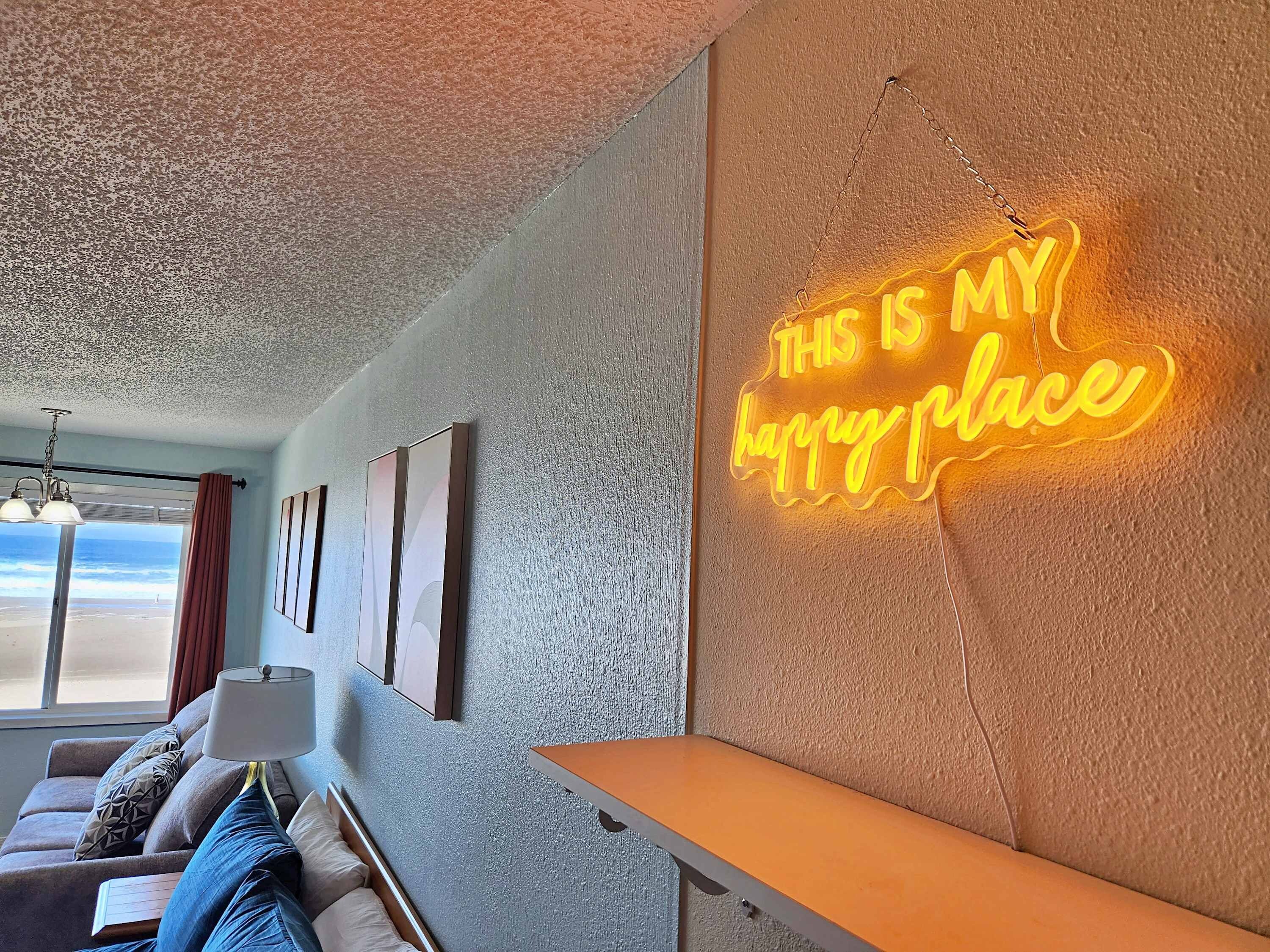 A living room with a neon sign on the wall, a sofa, a lamp, and a view of the beach.