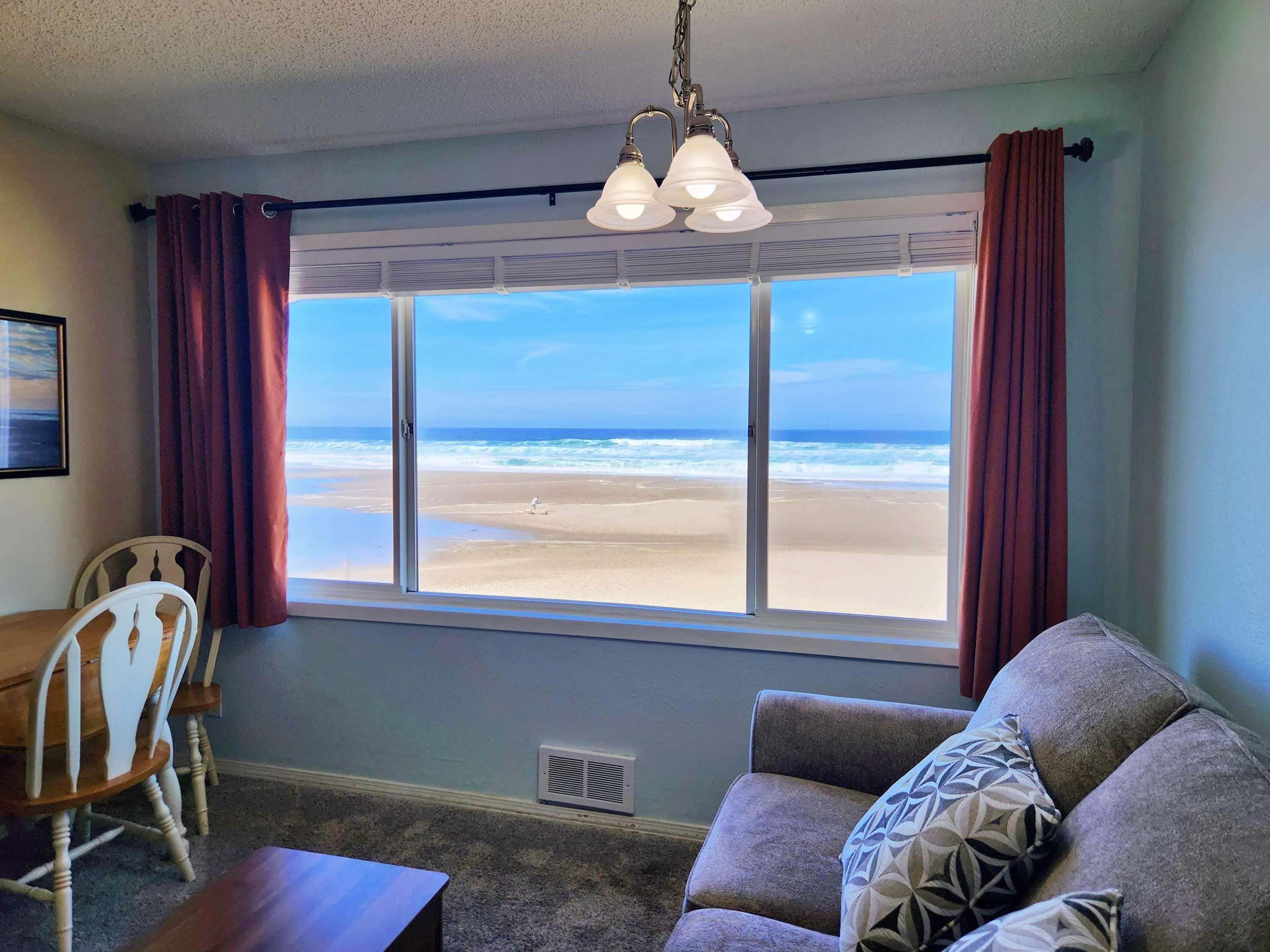 A room with a large window overlooking a beach, a dining table with chairs, and a sofa with patterned cushions.
