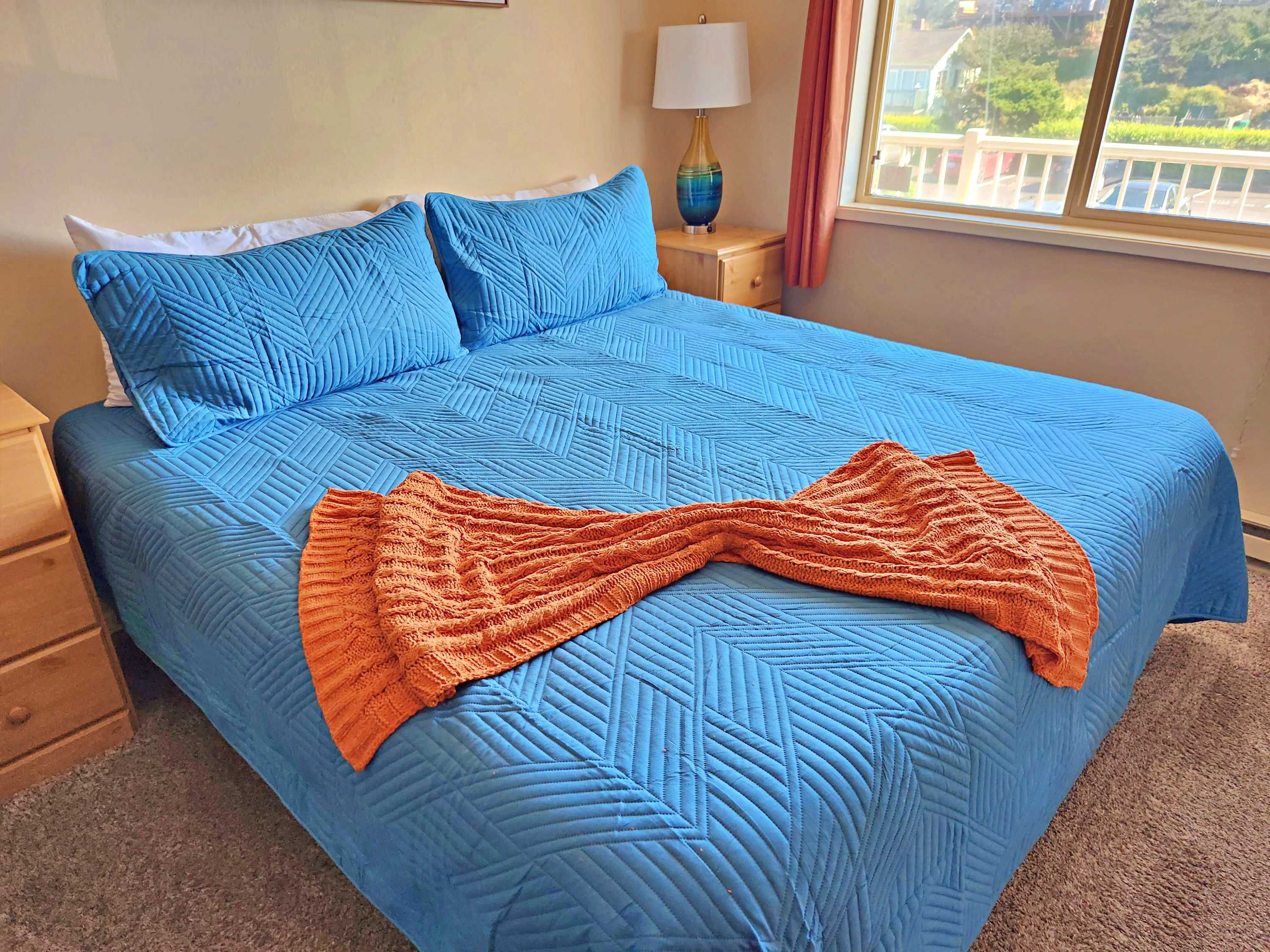 A bed with a blue quilt and pillows, a wooden nightstand with a lamp, and a window with a view of greenery.