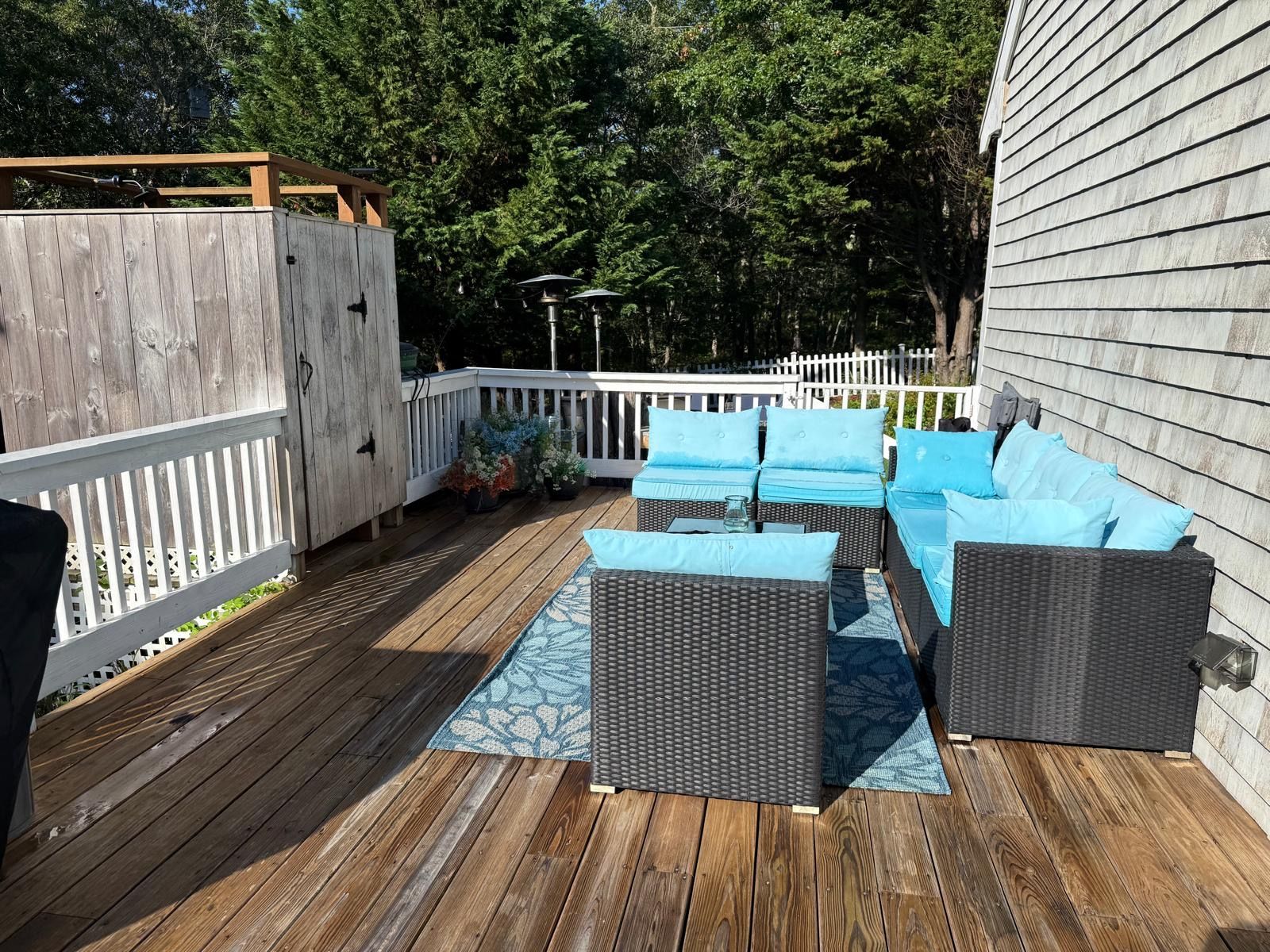 Een terras buiten met een houten vloer, een wit hek en een zithoek met blauwe kussens.
