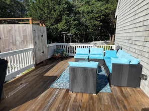 Een terras buiten met een houten vloer, een wit hek en een zithoek met blauwe kussens.
