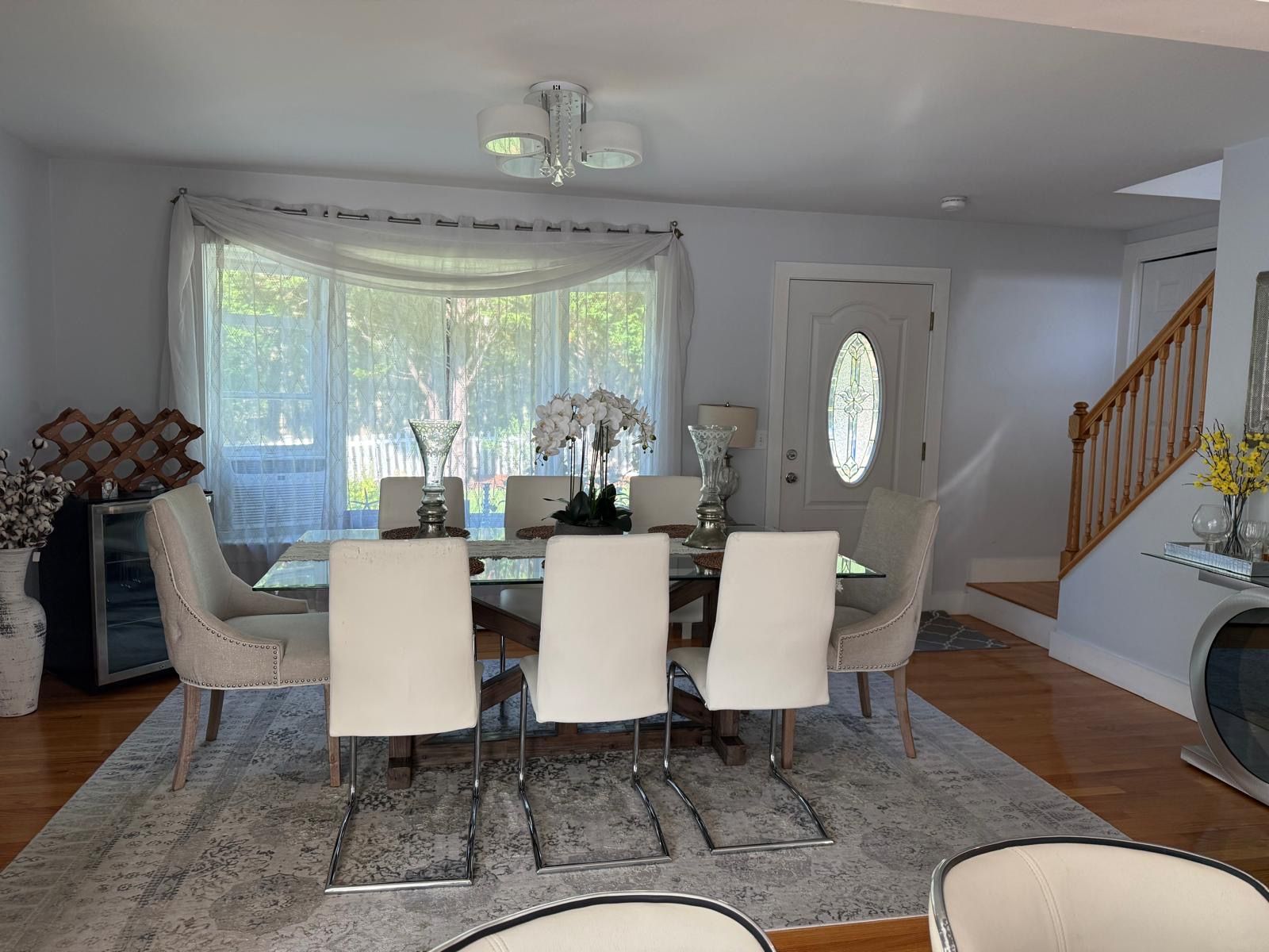A dining area with a table set for four, surrounded by chairs, a chandelier, and a view through large windows.