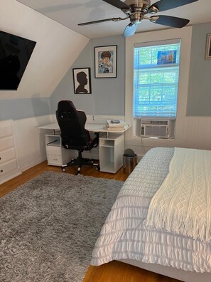 A bedroom with a bed, a desk, a chair, a ceiling fan, and a window with blinds.