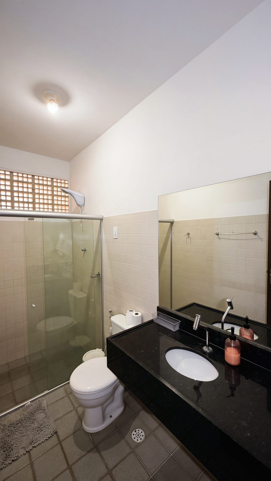 Un baño moderno con ducha de vidrio, inodoro blanco, lavamanos negro y paredes de azulejos color beige.