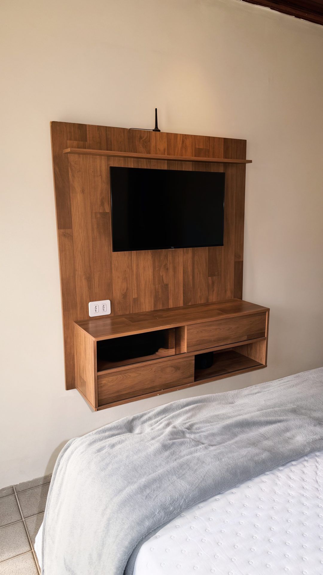 A wooden entertainment unit with a flat-screen TV mounted above it, a power outlet, and a lower shelf with drawers.