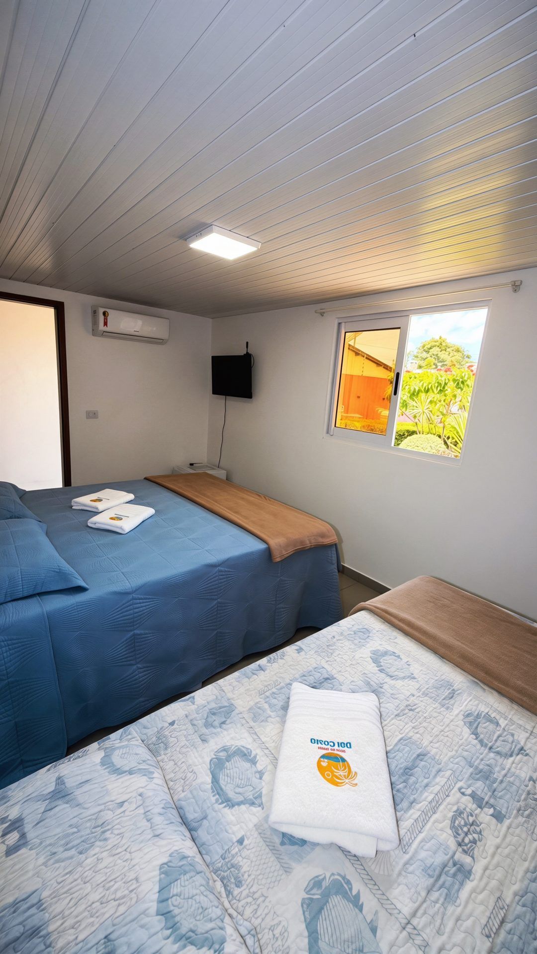A room with two beds, a flat-screen TV, an air conditioning unit, and a window with a view of greenery.