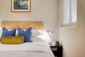Un lit bien fait, avec une tête de lit en osier, une lampe de chevet et un tableau encadré accroché au mur.
