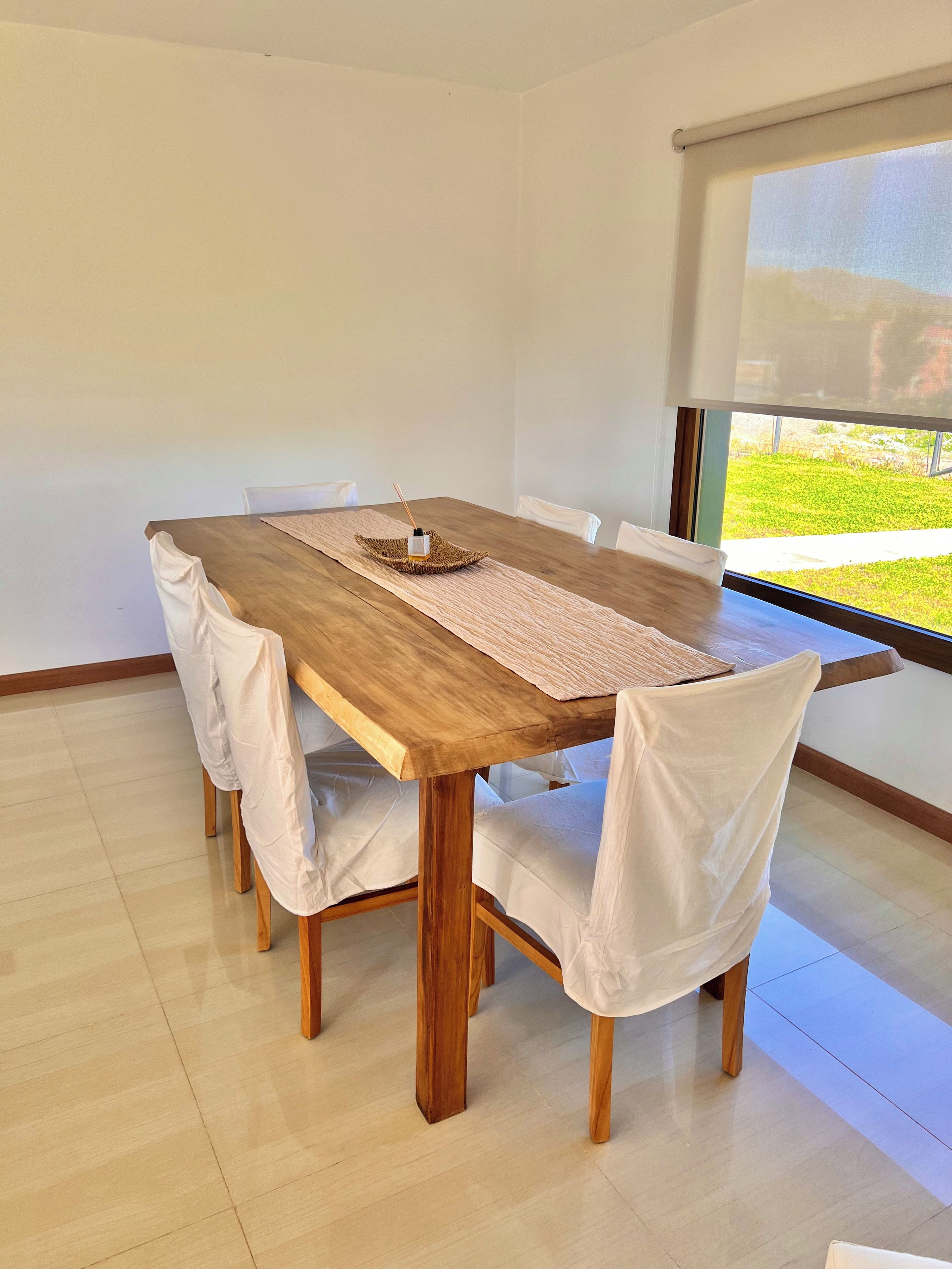 A dining area with a wooden table, white chairs, and a view of greenery outside.