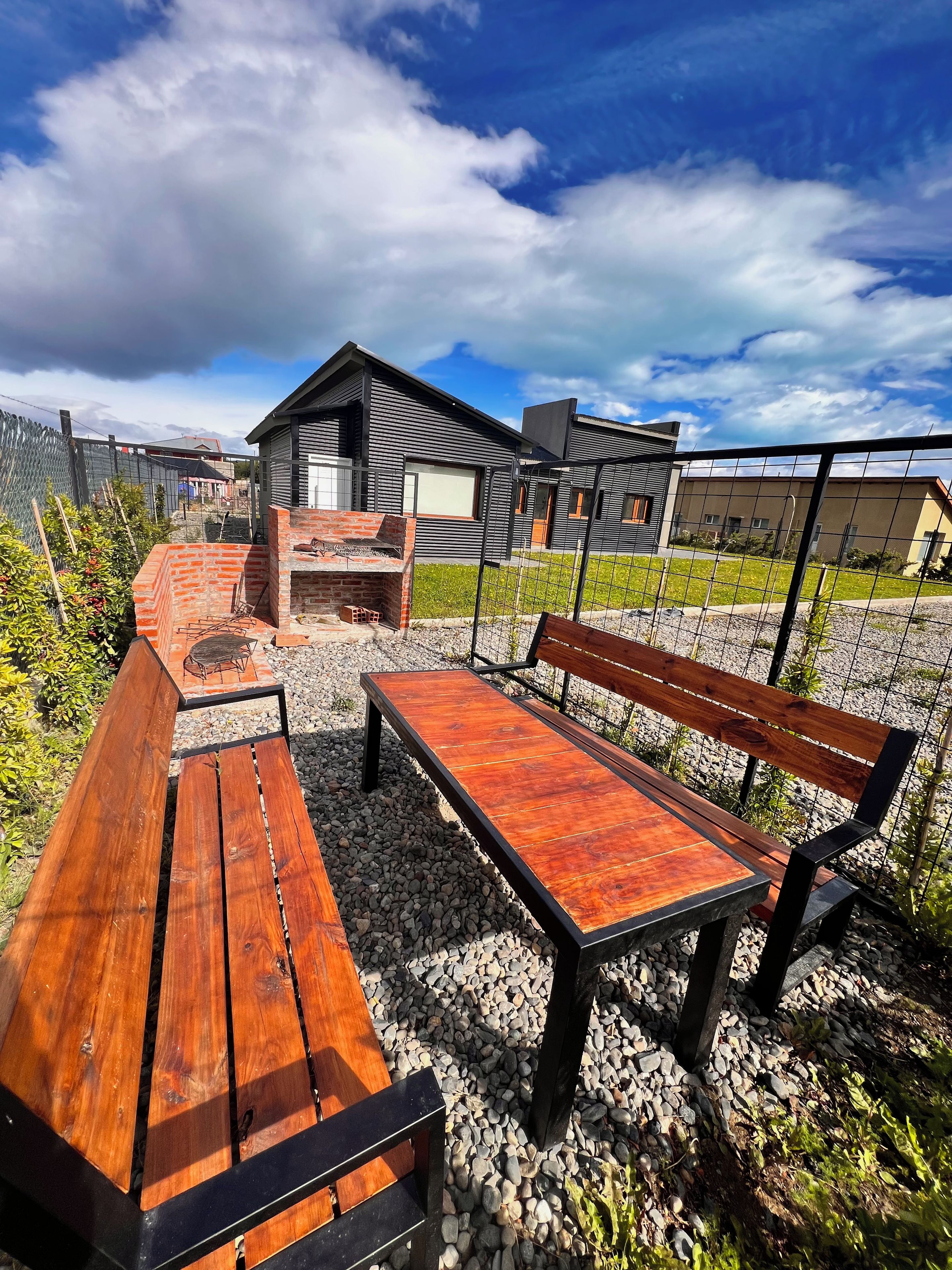An outdoor seating area with a wooden bench and table, surrounded by gravel and a metal fence.