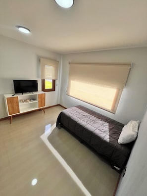 Una habitación compacta con cama, televisor sobre un mueble, ventana con persianas y una lámpara en el techo.