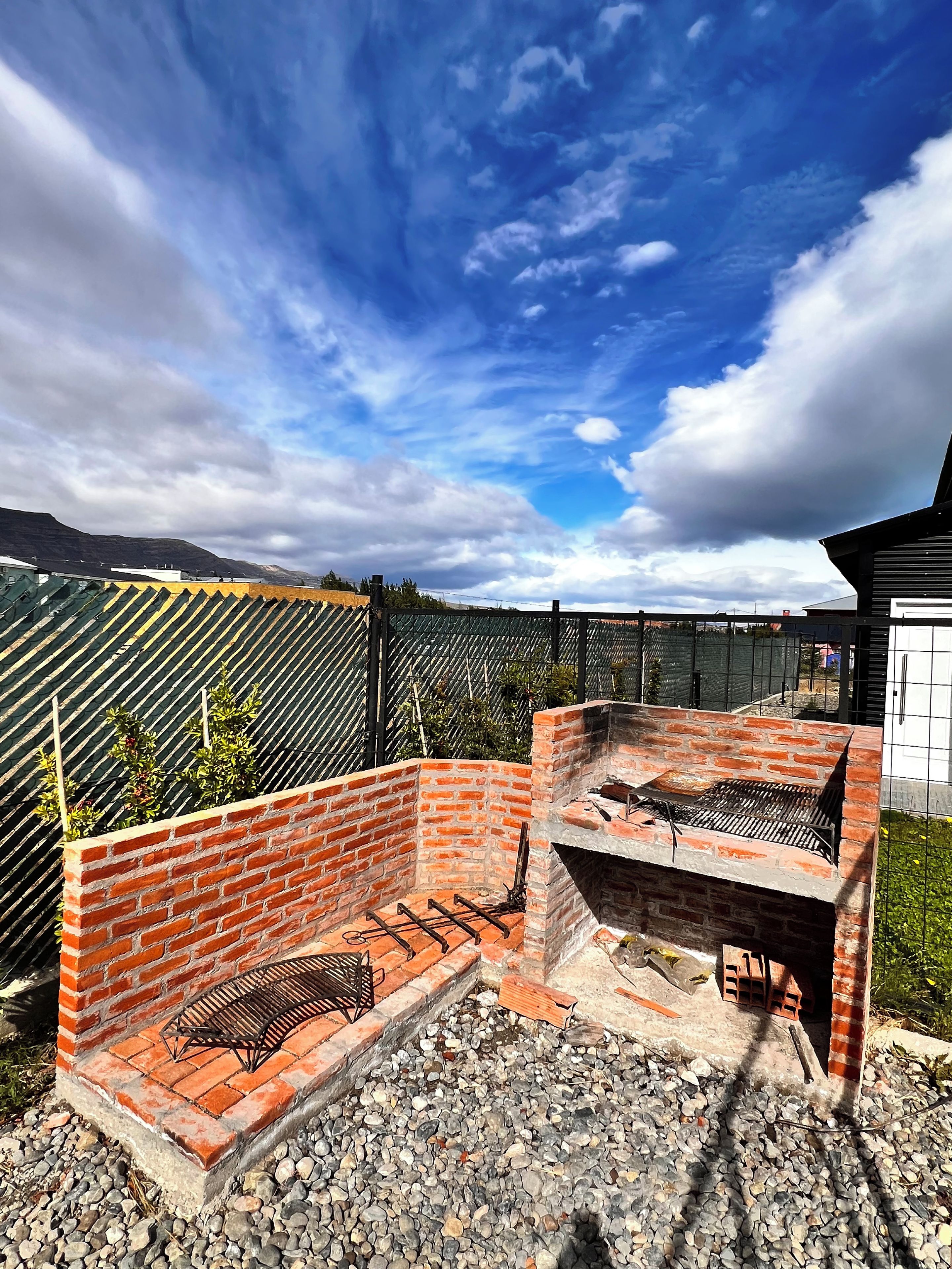 An outdoor barbecue area with brick walls and a grill, surrounded by a fence and a clear sky.