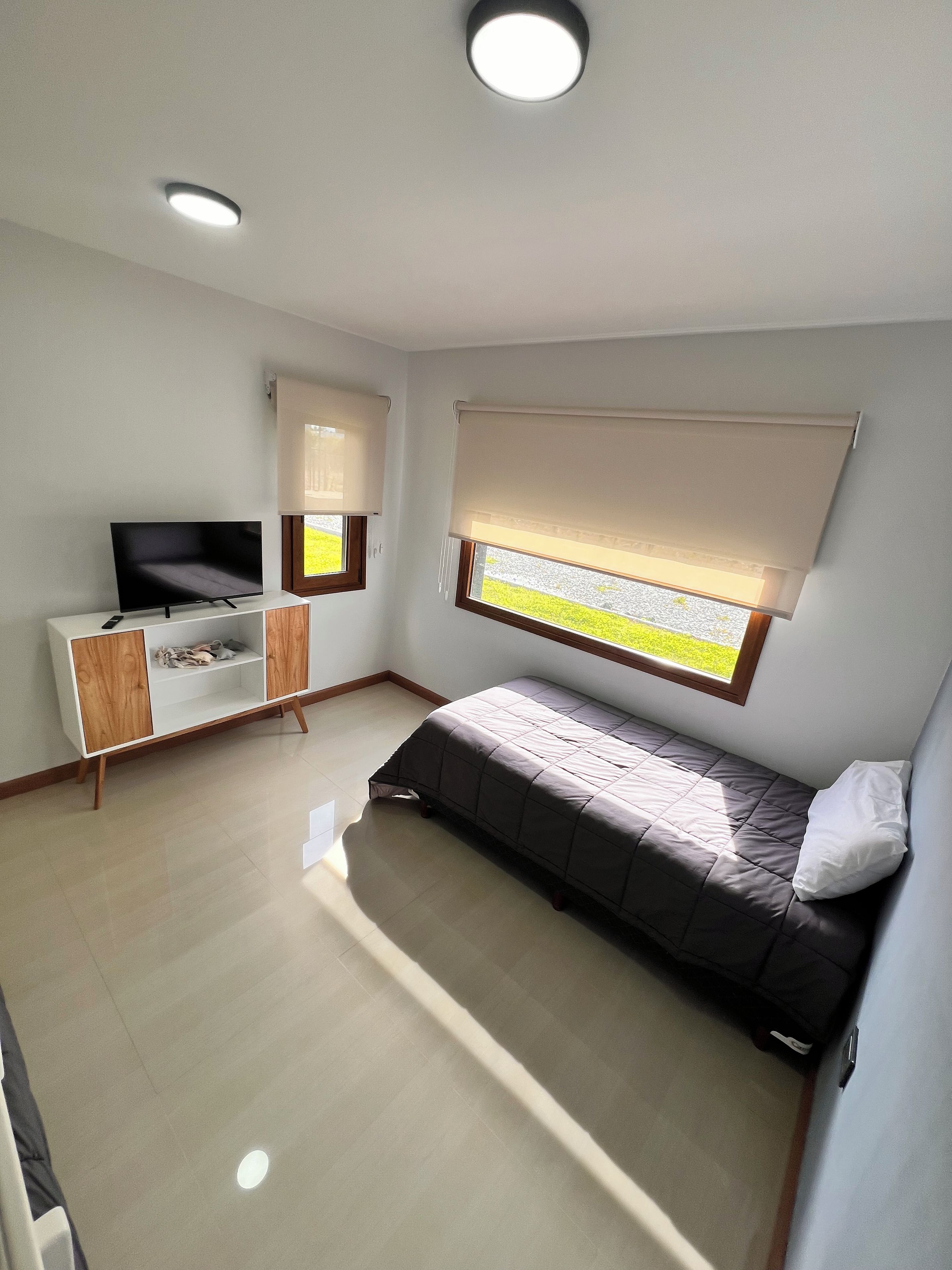 Een slaapkamer met een bed, een tv op een standaard, een raam met zonwering en een plafondlamp.