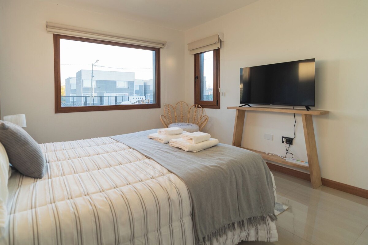 A bedroom with a bed, a television on a stand, a window, and a view of buildings outside.