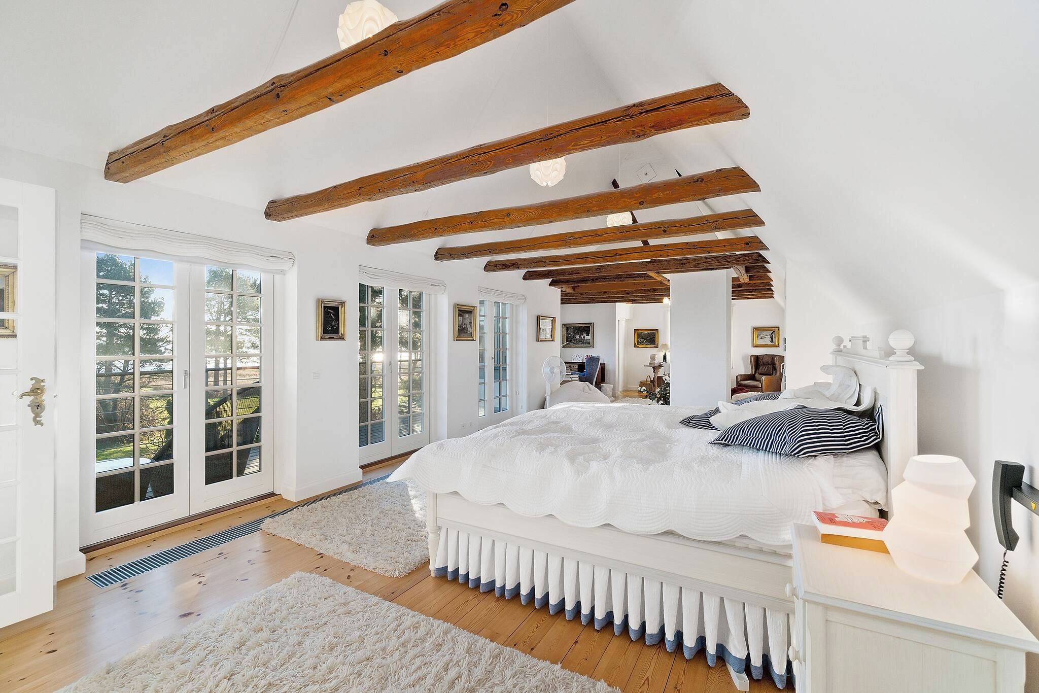 A bedroom with a large bed, wooden beams, white walls, and multiple windows.