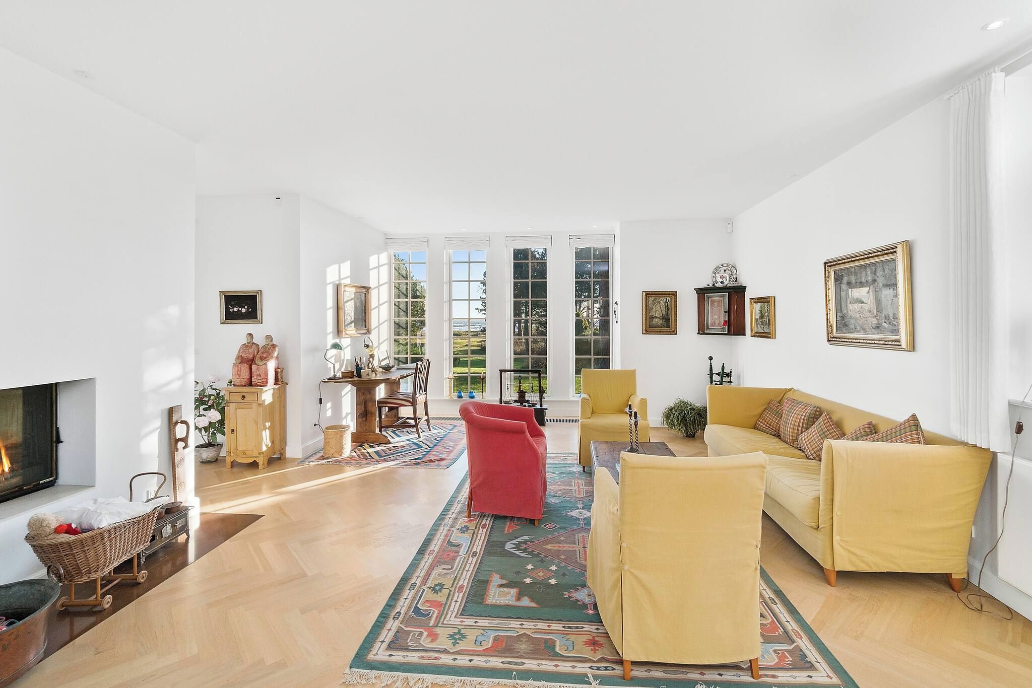 A spacious living room with a fireplace, a yellow sofa, a red chair, and a large window with a view of greenery.