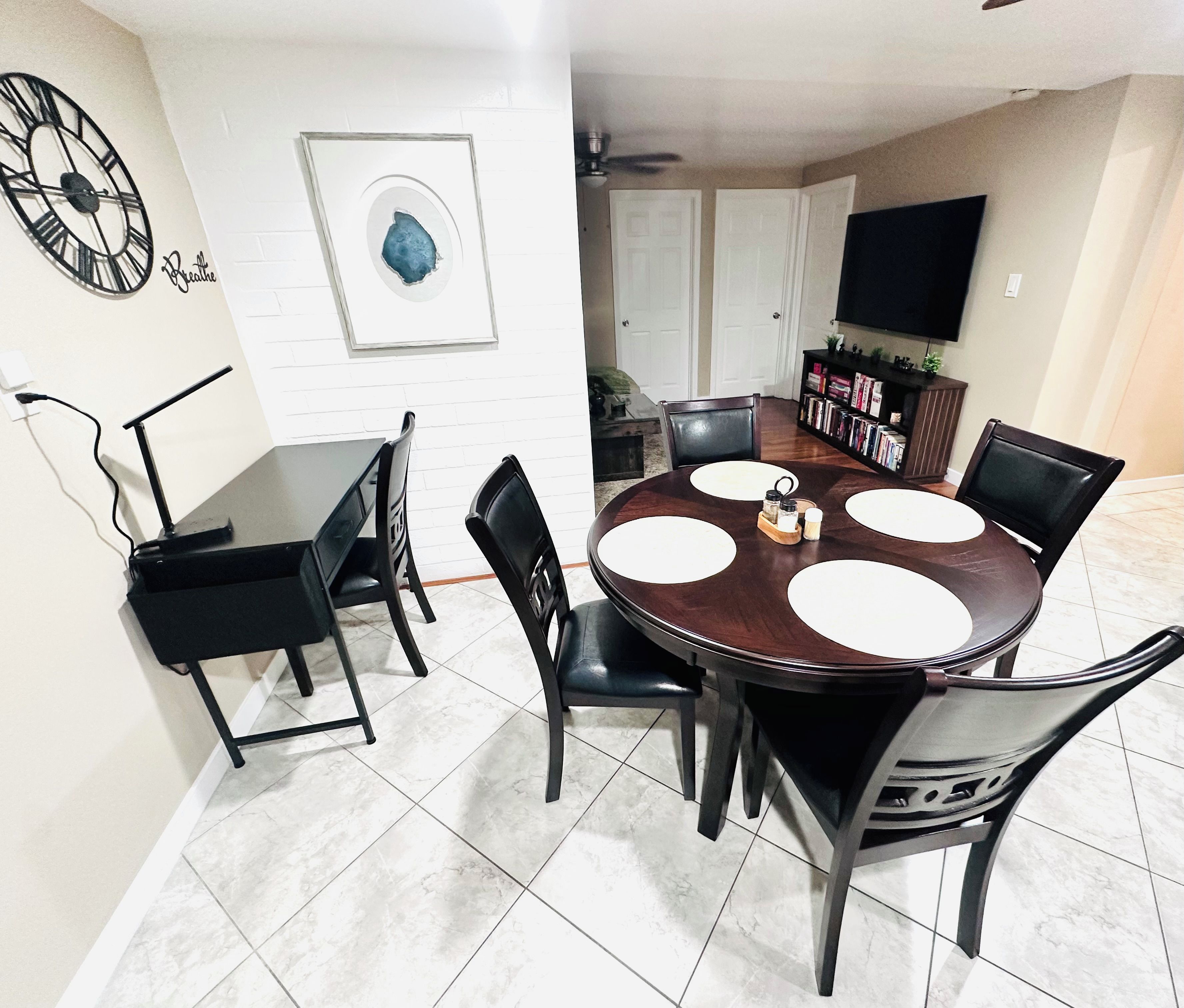 A dining area with a round table, black chairs, a clock, and a framed artwork on the wall.