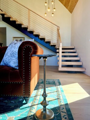 Een moderne woonkamer met een rode velourse bank, een kussen met dessin, een hoge vloerlamp, een blauw gekleurd vloerkleed en een trap met houten treden.