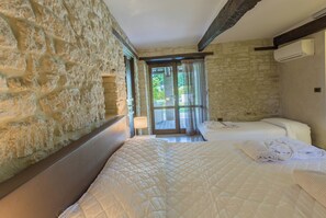 Quarto com parede de pedra, cama com colcha, mesa de cabeceira e janela com cortinas.