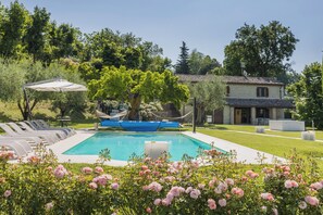 Área de piscina com espreguiçadeiras, uma rede e uma casa ao fundo.