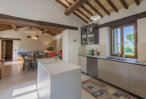 A modern kitchen with a central island, a dining area with a wooden table and chairs, and a view of the countryside through a window. - VILLA ANNA: HEATED POOL, 2 OUTDOOR HOT TUBS, SAUNA, AND GYM (Sant'Ippolito)