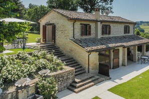 A stone house with a tiled roof, a covered porch, and a garden with white flowers. - VILLA ANNA: HEATED POOL, 2 OUTDOOR HOT TUBS, SAUNA, AND GYM (Sant'Ippolito)
