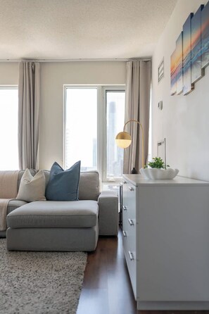 A modern living room with a sofa, a side table, a lamp, and a view of the city through large windows. - Luxury Downtown Toronto Condo with Stunning City Views | Parking Available (Toronto)