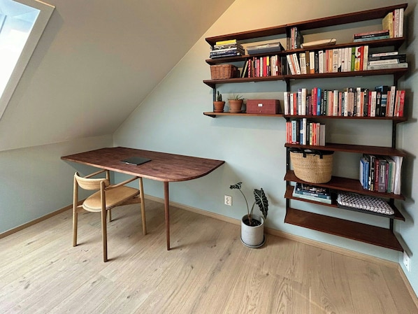 Ein Zimmer mit schrägem Dach, einem Holztisch mit Stuhl und einem Bücherregal voller Bücher.