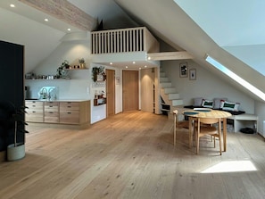 Ein geräumiges Loft mit hoher Decke, Holzboden, einer Essecke mit Tisch und Stühlen sowie einer Küche mit Holzschränken und einer Spüle.