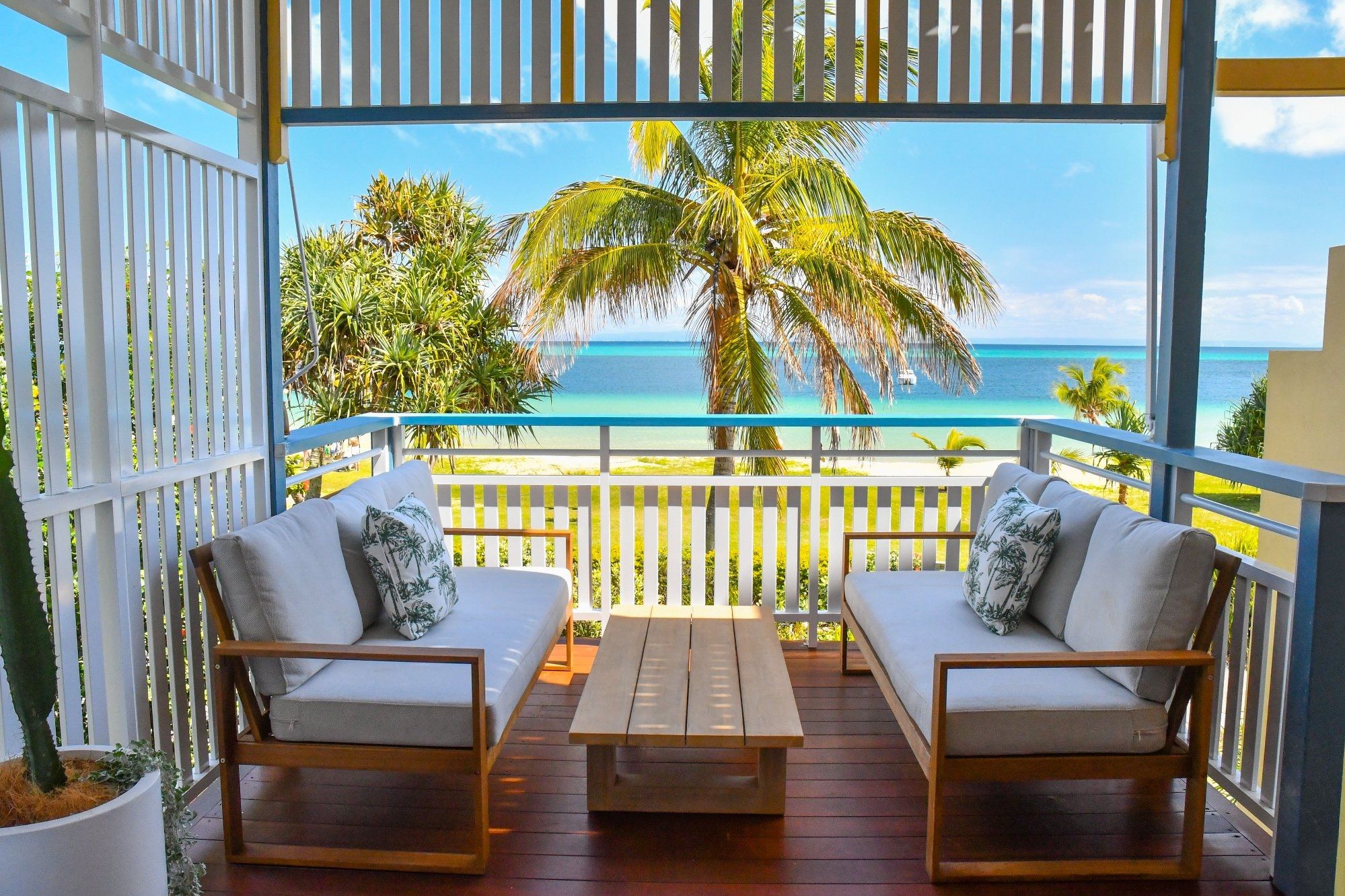 A balcony with two sofas and a table, overlooking a beach with palm trees and the ocean.