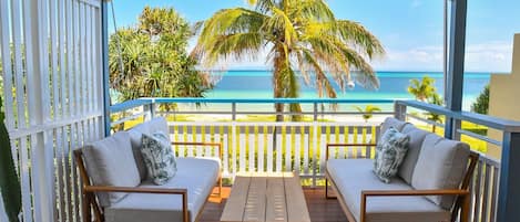A balcony with two sofas and a table, overlooking a beach with palm trees and the ocean.