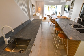A modern kitchen with a central island, bar stools, and a dining area with a view to the outdoors. - Villa 33 - Tangalooma - Moreton Island (moreton island)