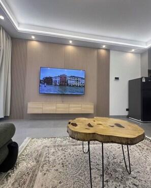 A modern living room with a flat-screen TV mounted on a wooden wall, a round wooden coffee table with metal legs, and a patterned rug on the floor. - Chic 2BR with HeatedPool, Gym, Sauna, SteamRoom, Wi-fi , Safe Parking (Nairobi)