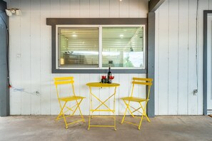 Un petit espace extérieur aménagé avec deux chaises jaunes et une table, situé sous une fenêtre munie de stores.