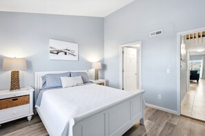 Un dormitorio con cama, mesitas de noche, lámpara, un cuadro en la pared y una puerta que da a otra habitación.