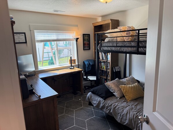 A dormitory room with bunk beds, a desk with a computer, a bookshelf, and a window with blinds.