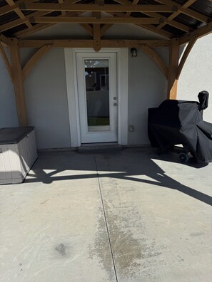 A covered patio area with a white door, a barbecue grill, and a concrete floor.