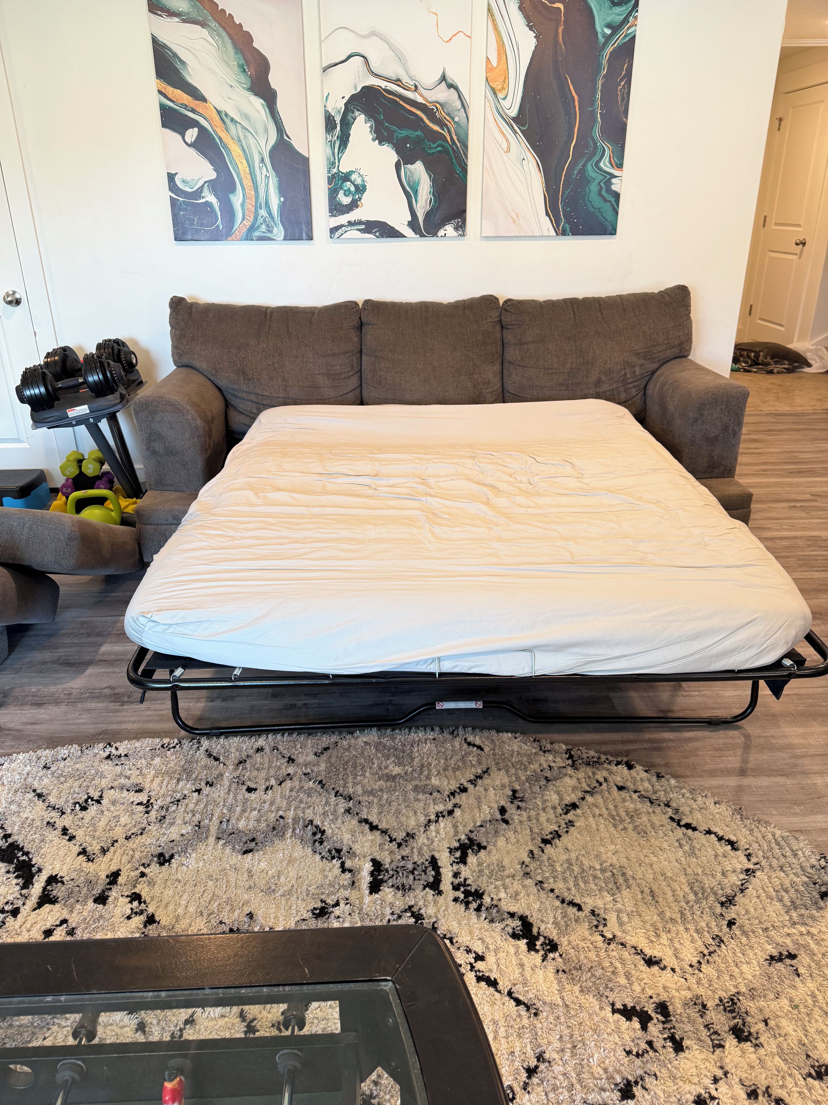 Un lit avec un matelas, un canapé gris, une petite table avec des haltères et des œuvres d’art abstraites accrochées au mur.