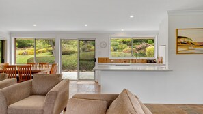 A modern kitchen with a large window overlooking a garden, a dining area with wooden furniture, and a living space with a sofa. - Bawley Point Coastal Acreage - Guest Retreat (Bawley Point)