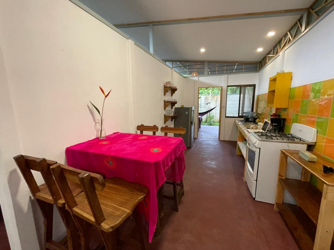 A dining area with a table covered in a bright pink tablecloth, wooden chairs, and a vase with flowers. A kitchen with a stove and cabinets is visible in the background.