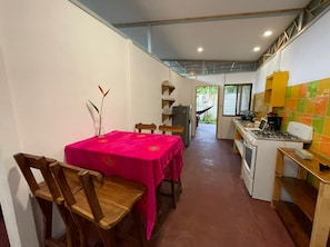 A dining area with a table covered in a bright pink tablecloth, wooden chairs, and a vase with flowers. A kitchen with a stove and cabinets is visible in the background.