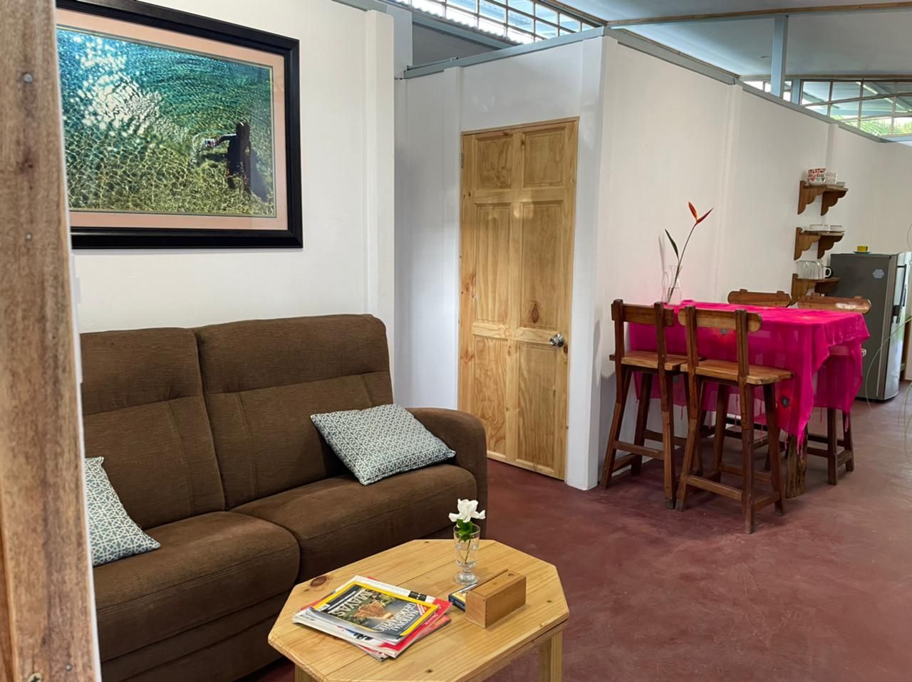 A living room with a brown sofa, a wooden coffee table with magazines, a wooden dining table with a pink tablecloth, and a wooden door.