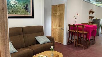 A living room with a brown sofa, a wooden coffee table with magazines, a wooden dining table with a pink tablecloth, and a wooden door.