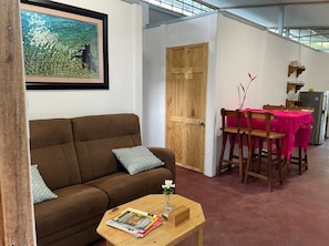 A living room with a brown sofa, a wooden coffee table with magazines, a wooden dining table with a pink tablecloth, and a wooden door.