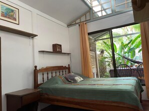 A bedroom with a bed, a nightstand, a shelf with a picture, and a window with curtains.