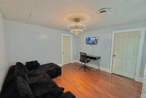 Ein Zimmer mit einer schwarzen Sofagarnitur, einem Schreibtisch mit Stuhl, einem an der Wand montierten Fernseher und zwei Türen.