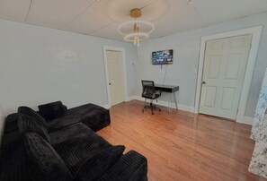 Ein Zimmer mit einer schwarzen Sofagarnitur, einem Schreibtisch mit Stuhl, einem Fernseher an der Wand und einer Tür.